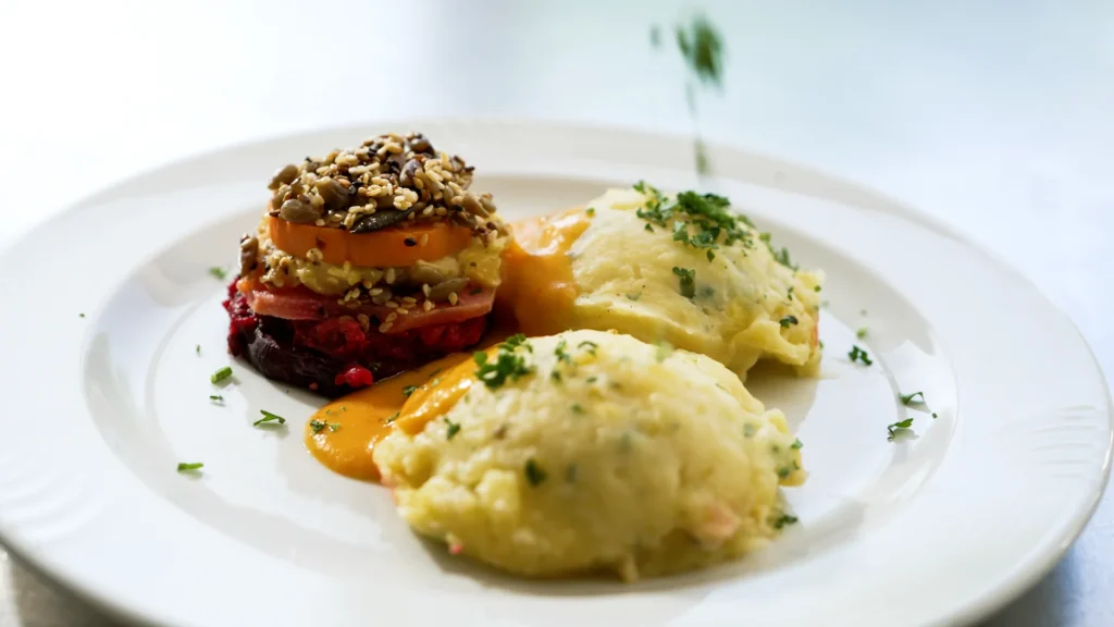 Auf einem weißen Teller liegen ein Gemüsetürmchen, zwei Portionen Püree und eine Garnitur aus Kräutern.