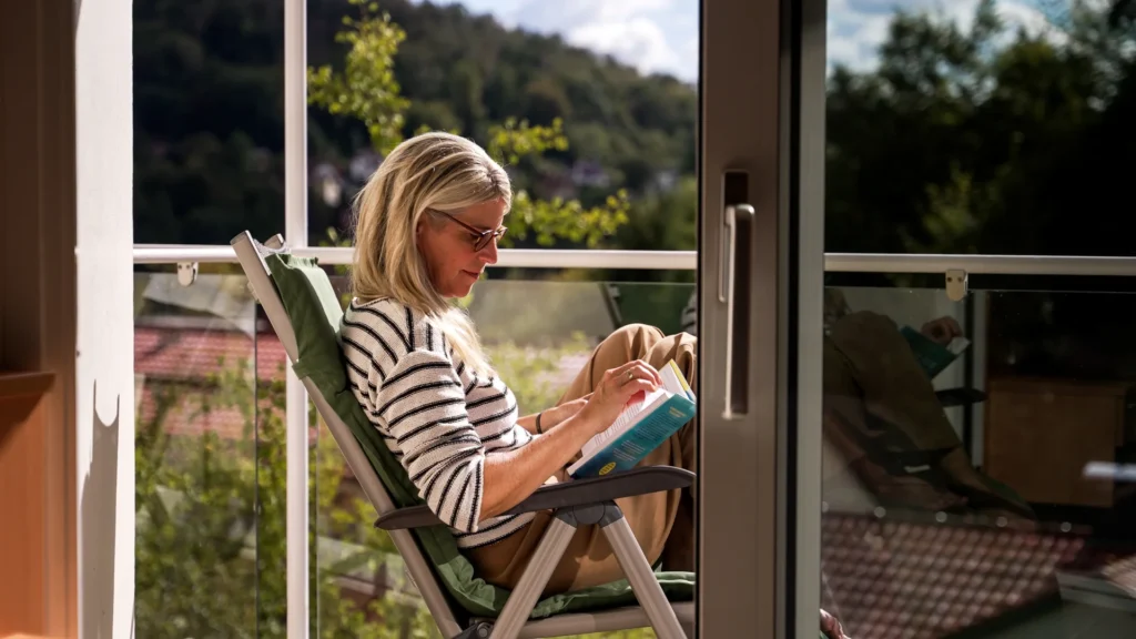 Eine Frau sitzt auf dem sonnigen Balkon in einem Klappstuhl und liest in einer aufgeschlagenen Zeitschrift.