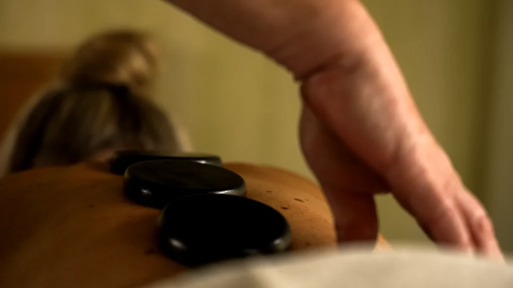 Dunkle Massagesteine liegen auf einem Rücken, während eine Hand die Schulter der liegenden Person berührt.