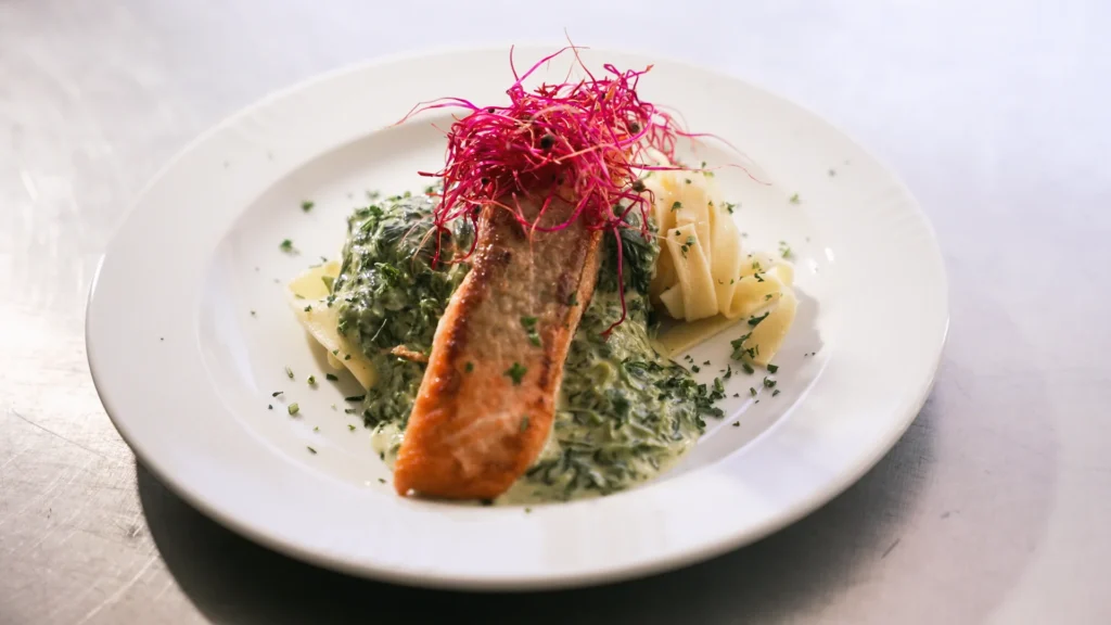 Ein Lachsfilet wird auf einem weißen Teller mit Bandnudeln, grüner Sauce und roten Sprossen serviert.