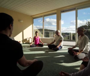 Mehrere Personen sitzen im Kreis auf Matten im hellen Meditationsraum mit großen Fenstern und Balkontür. Mehrere Personen sitzen im Kreis auf Matten im hellen Meditationsraum mit großen Fenstern und Balkontür.