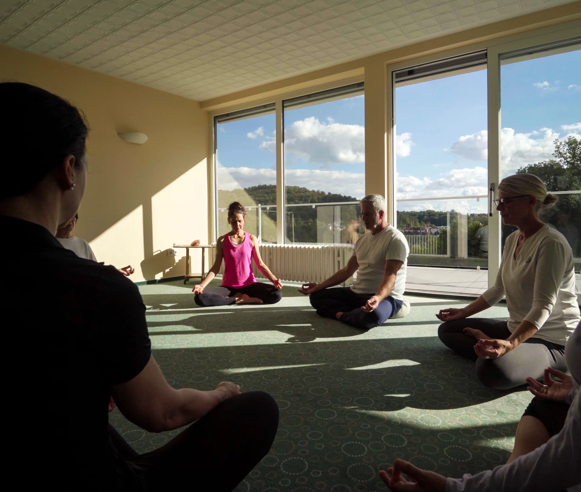 Mehrere Personen sitzen im Kreis auf Matten im hellen Meditationsraum mit großen Fenstern und Balkontür.
