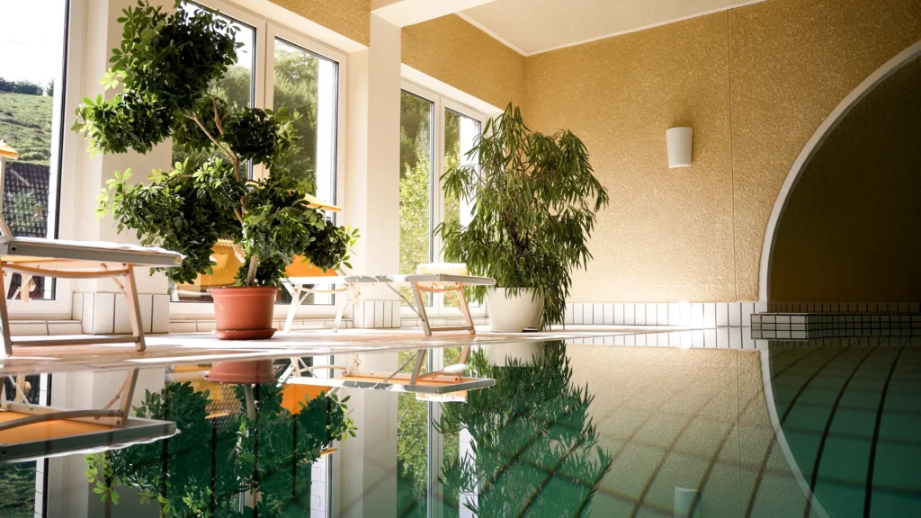 Sonnenlicht und große Zimmerpflanzen spiegeln sich im ruhigen Wasser des Schwimmbeckens vor der Fensterfront.