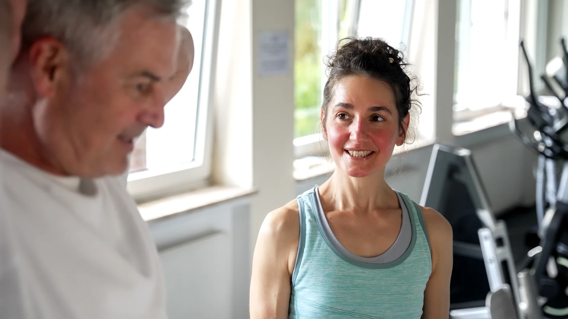 Unsere Physiotherapeutin steht lächelnd neben einem Gast im Trainingsraum mit großen Fenstern.