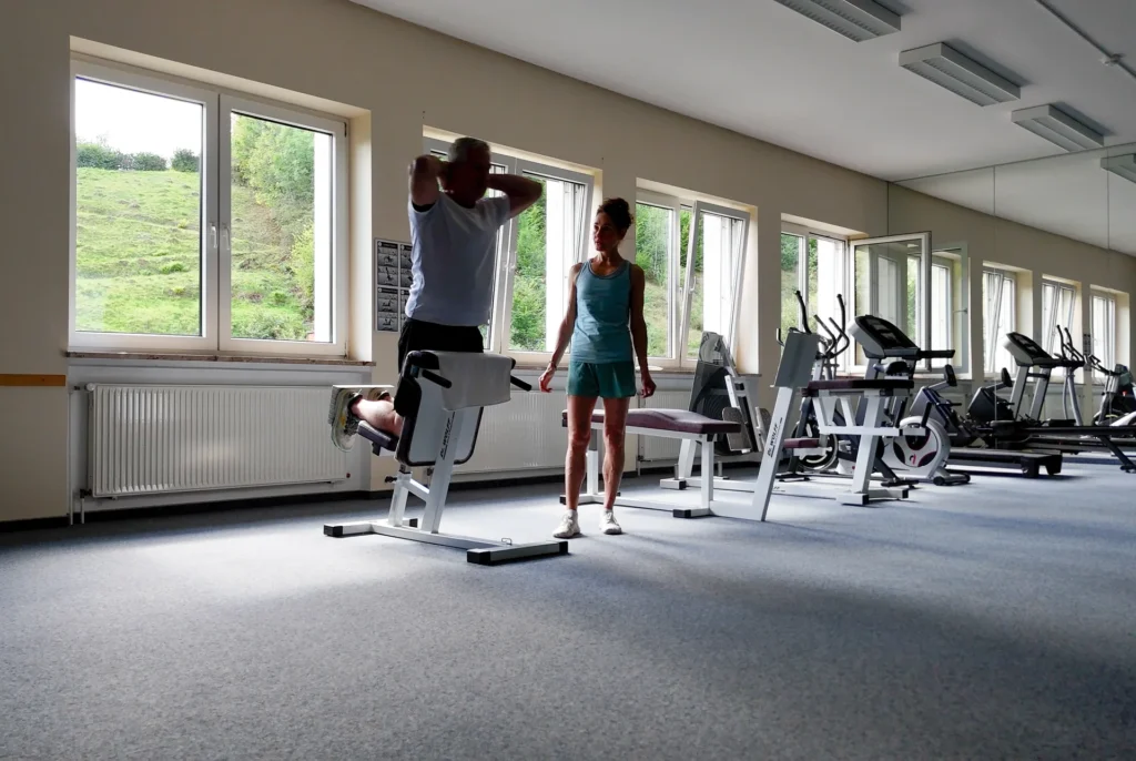 Ein Mann trainiert an einem Gerät, während eine Physiotherapeutin danebensteht; weitere Fitnessgeräte stehen am Fenster. Ein Mann trainiert an einem Gerät, während eine Physiotherapeutin danebensteht; weitere Fitnessgeräte stehen am Fenster.