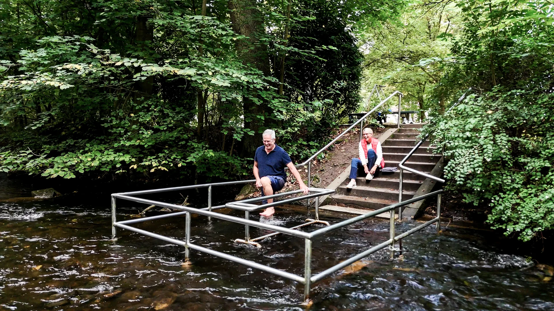 Zwei Personen nutzen die Wassertretstelle im Kurpark Bad Lauterberg mit Metallgeländern im fließenden Wasser.